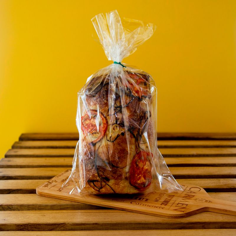 Pain artisanal à la tomate emballé sous film plastique sur une planche en bois rustique, fond jaune.