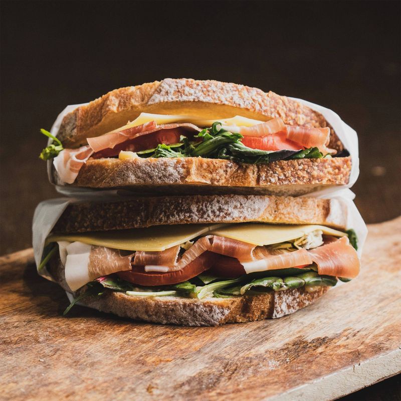 Gros plan sur un sandwich gastronomique avec jambon cru, fromage et verdure sur une planche en bois.